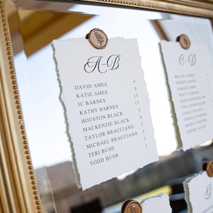 May include: Elegant wedding seating chart with names and table numbers printed on textured paper. Each card is held by a decorative gold-colored seal with a leaf design. The cards are displayed in a gold-framed mirror.