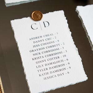 May include: Elegant wedding place cards with torn edges, featuring names and table numbers. Each card is topped with a gold wax seal. The cards are arranged on a reflective surface, creating a sophisticated display.