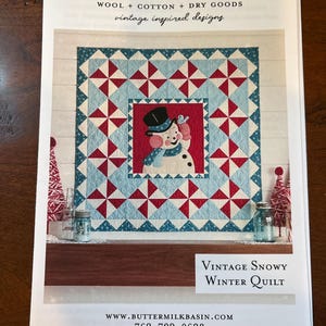 May include: A printed pattern sheet for a quilt titled Vintage Snowy Winter Quilt by Buttermilk Basin. The quilt features a central snowman wearing a black top hat and blue scarf, surrounded by a red, light blue, and white pinwheel border pattern.