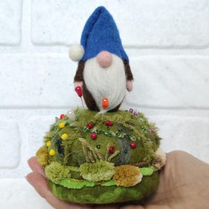 Puede incluir: Un gnomo de fieltro con un sombrero azul y barba blanca se asienta sobre un alfiletero verde texturizado. La base est&aacute; decorada con peque&ntilde;os mechones de fibras verdes y marrones, con varios alfileres de cabezas redondas de colores.