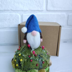 Puede incluir: Un gnomo de lana fieltrada con sombrero azul y barba blanca se asienta sobre un acerico redondo de color verde musgo. El acerico est&aacute; decorado con peque&ntilde;as cuentas verdes y amarillas, parches de tela texturizada y alfileres rojos.