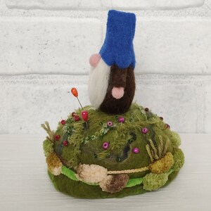 Puede incluir: Un gnomo de lana fieltrada con sombrero azul, barba blanca y cuerpo marr&oacute;n se asienta sobre una base de alfiletero verde que imita el musgo. La base est&aacute; decorada con peque&ntilde;as cuentas de colores, mechones de hilo y dos alfileres verticales.