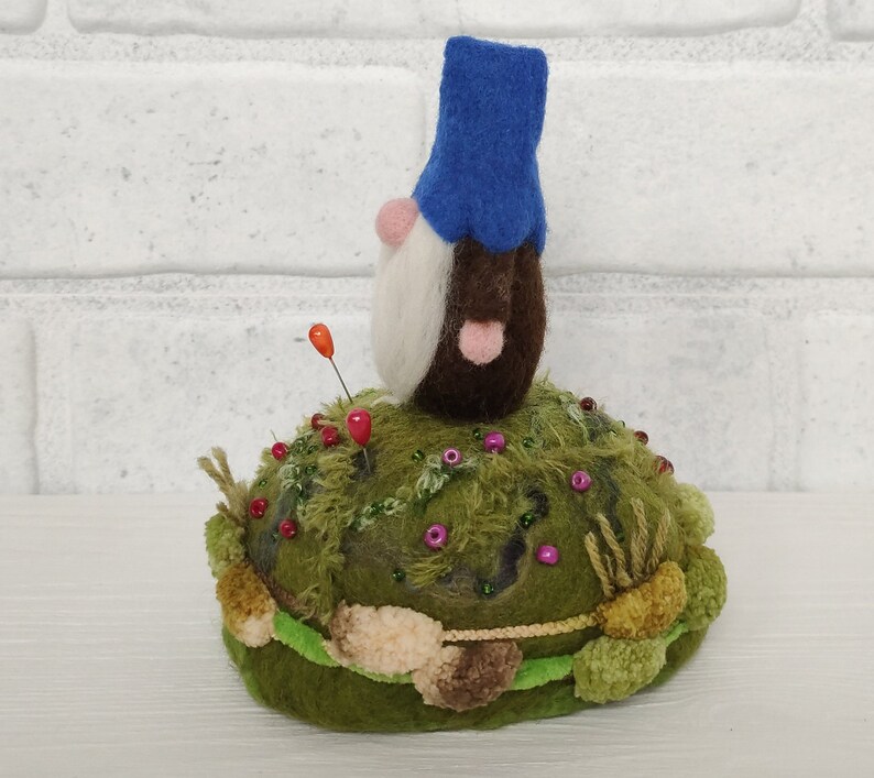 Puede incluir: Un gnomo de lana fieltrada con sombrero azul, barba blanca y cuerpo marr&oacute;n se asienta sobre una base de alfiletero verde que imita el musgo. La base est&aacute; decorada con peque&ntilde;as cuentas de colores, mechones de hilo y dos alfileres verticales.