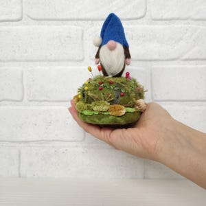 Puede incluir: Un gnomo de lana fieltrada con sombrero azul y barba blanca sobre un acerico redondo de color verde. La base est&aacute; decorada con musgo verde texturizado, peque&ntilde;os pompones marrones y varios alfileres de colores con cabeza redonda.