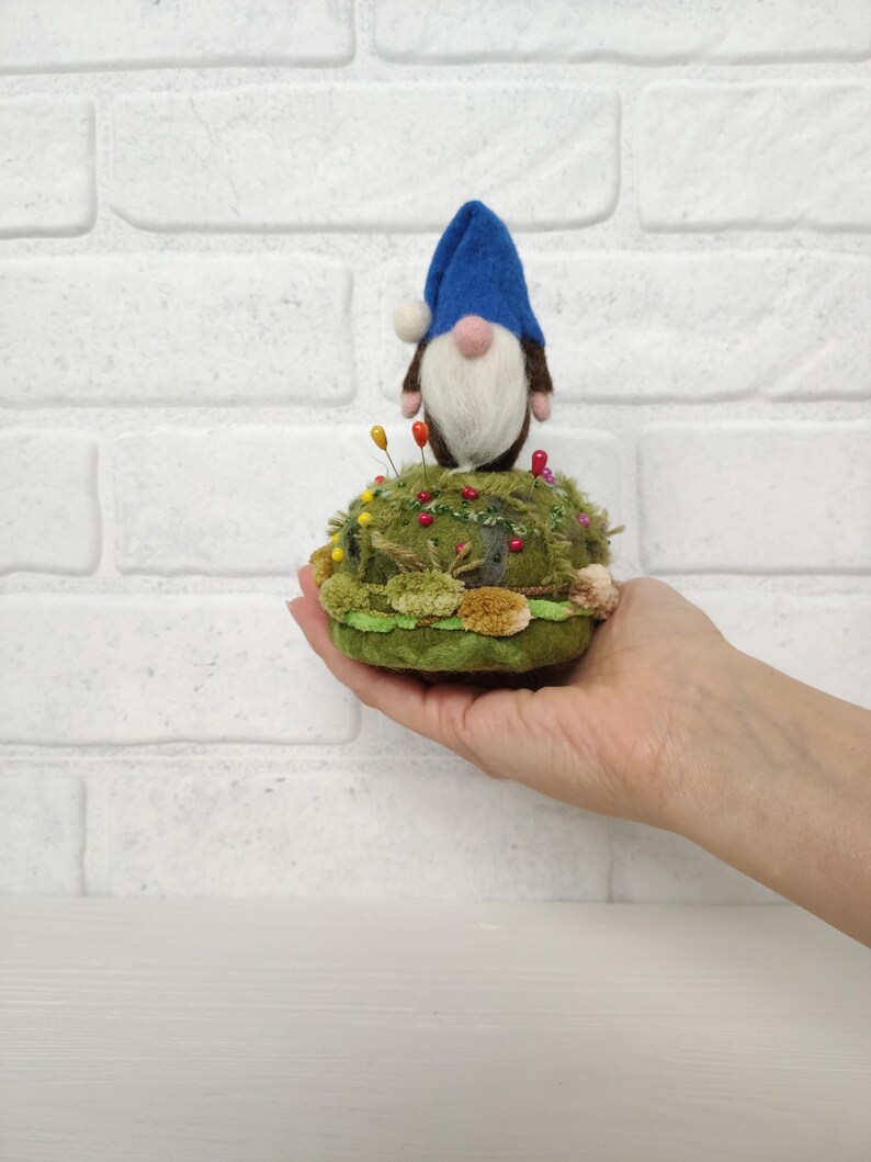 Puede incluir: Un gnomo de lana fieltrada con sombrero azul y barba blanca sobre un acerico redondo de color verde. La base est&aacute; decorada con musgo verde texturizado, peque&ntilde;os pompones marrones y varios alfileres de colores con cabeza redonda.