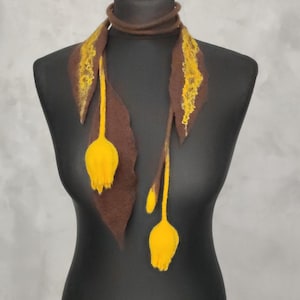 Puede incluir: Una bufanda de fieltro marrón con dos extremos largos en forma de hoja con detalles florales texturizados en amarillo brillante. La bufanda se envuelve alrededor del cuello con una banda fina y tiene dos formas florales amarillas colgantes.
