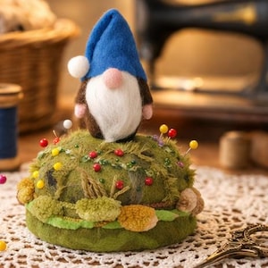Puede incluir: Un acerico con una base redonda de color verde musgo y textura, adornado con peque&ntilde;os pompones coloridos y m&uacute;ltiples alfileres con cabezas rojas, amarillas y blancas. Un peque&ntilde;o gnomo de fieltro con gorro azul y barba blanca se asienta encima.