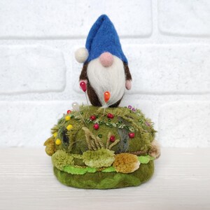 Puede incluir: Una figura de gnomo de lana fieltrada con un sombrero azul y barba blanca, colocada sobre una base de alfiletero redonda y texturizada de color verde musgo. La base est&aacute; decorada con peque&ntilde;as cuentas coloridas y varias texturas de tela verde, sirviendo como soporte para varios alfileres de costura con cabezas rojas, amarillas y blancas.