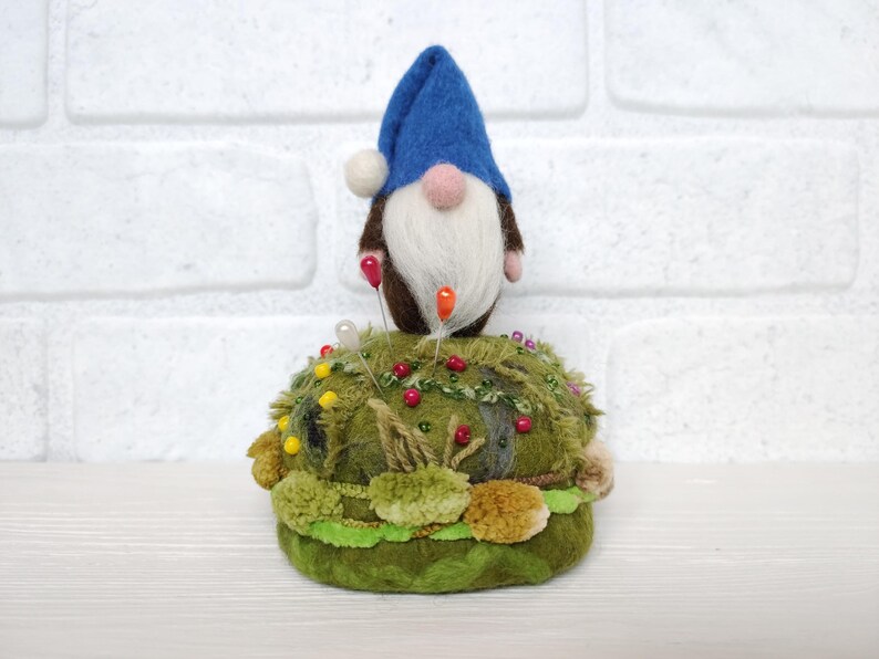 Puede incluir: Una figura de gnomo de lana fieltrada con un sombrero azul y barba blanca, colocada sobre una base de alfiletero redonda y texturizada de color verde musgo. La base est&aacute; decorada con peque&ntilde;as cuentas coloridas y varias texturas de tela verde, sirviendo como soporte para varios alfileres de costura con cabezas rojas, amarillas y blancas.