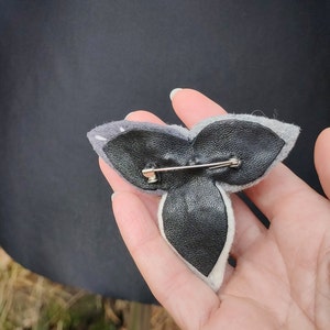 以下が含まれることがあります： グレーと白のフェルト製のブローチ。裏側は黒革で、安全ピンで留めるタイプです。