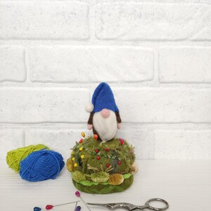 Puede incluir: Un gnomo de fieltro con sombrero azul y barba blanca se asienta sobre un acerico redondo de color verde musgo. A su lado hay dos peque&ntilde;as bolas de lana verde y azul, varios alfileres de colores y unas tijeras ornamentadas de metal plateado.