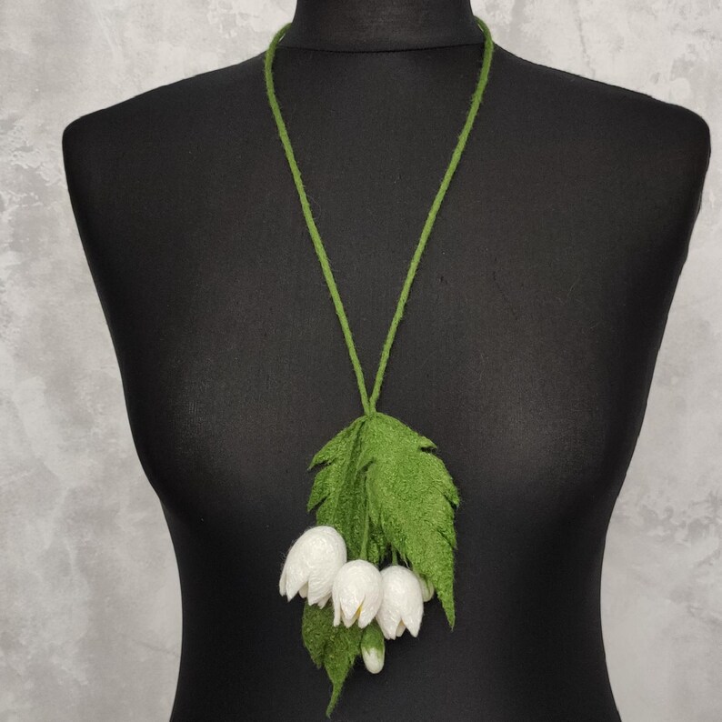 Pu&ograve; includere: Una collana di fiori in feltro bianco con foglie in feltro verde. La collana &egrave; su un manichino nero.