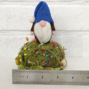 Puede incluir: Un gnomo de fieltro con sombrero azul y barba blanca se asienta sobre un acerico redondo de color verde musgo con textura, decorado con peque&ntilde;as cuentas y alfileres. Una regla met&aacute;lica muestra una base de unos 11,5 cm de ancho.