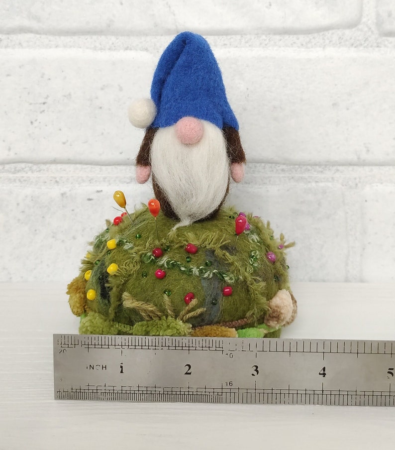Puede incluir: Un gnomo de fieltro con sombrero azul y barba blanca se asienta sobre un acerico redondo de color verde musgo con textura, decorado con peque&ntilde;as cuentas y alfileres. Una regla met&aacute;lica muestra una base de unos 11,5 cm de ancho.