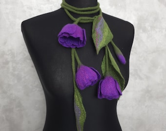 May include: A felted scarf featuring three purple tulip-like flowers with green stems and leaves. The scarf is displayed on a black mannequin, showcasing the floral design and color contrast. The scarf is a unique accessory.
