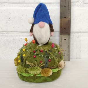 Puede incluir: Un gnomo de fieltro con un sombrero azul y barba blanca se asienta sobre un acerico redondo de color verde musgo, decorado con peque&ntilde;as cuentas de colores y texturas de hilo. Una regla met&aacute;lica muestra una altura de unos 9 cm.