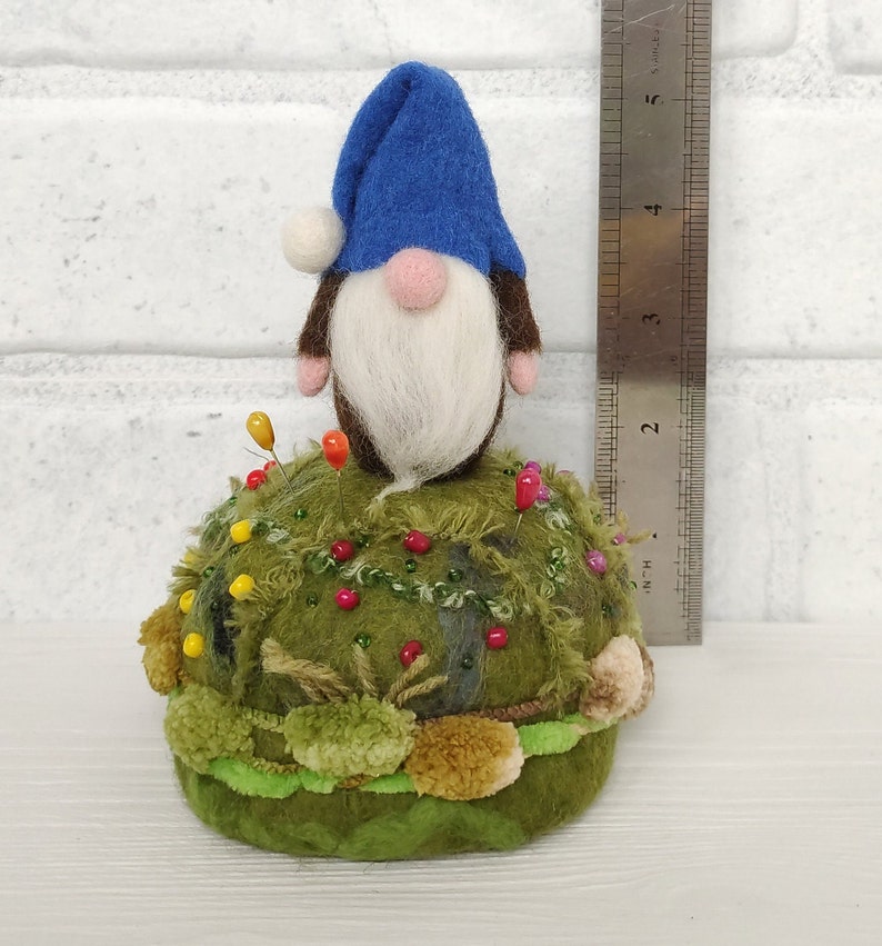 Puede incluir: Un gnomo de fieltro con un sombrero azul y barba blanca se asienta sobre un acerico redondo de color verde musgo, decorado con peque&ntilde;as cuentas de colores y texturas de hilo. Una regla met&aacute;lica muestra una altura de unos 9 cm.