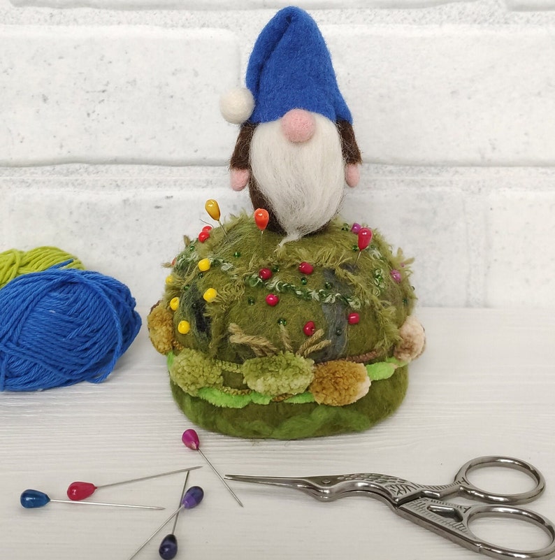 Puede incluir: Un gnomo de lana fieltrada con sombrero azul y barba blanca se asienta sobre un acerico redondo de color verde musgo, decorado con peque&ntilde;os pompones, cuentas y alfileres de colores. Junto al acerico hay ovillos de lana y tijeras.