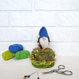 Puede incluir: Un gnomo de lana fieltrada con gorro azul y barba blanca se asienta sobre un acerico redondo verde decorado con texturas de musgo y alfileres de colores. Junto a &eacute;l hay dos ovillos de lana azul y verde, dos alfileres sueltos y unas tijeras.