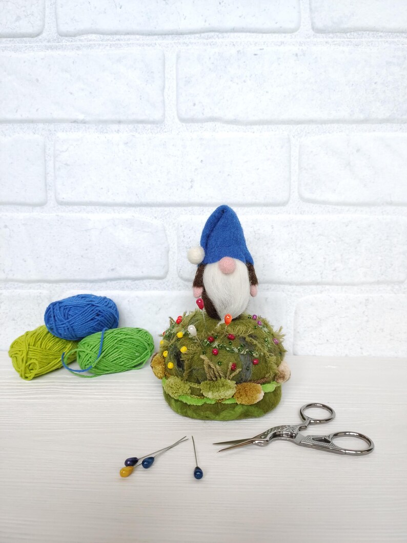 Puede incluir: Un gnomo de lana fieltrada con gorro azul y barba blanca se asienta sobre un acerico redondo verde decorado con texturas de musgo y alfileres de colores. Junto a &eacute;l hay dos ovillos de lana azul y verde, dos alfileres sueltos y unas tijeras.