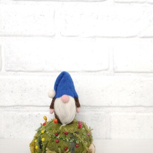 Puede incluir: Un gnomo de lana fieltrada con un gorro azul y barba blanca se asienta sobre un acerico redondo de color verde musgo. El acerico est&aacute; decorado con varios alfileres de colores, peque&ntilde;as cuentas y capas de tela texturizada.