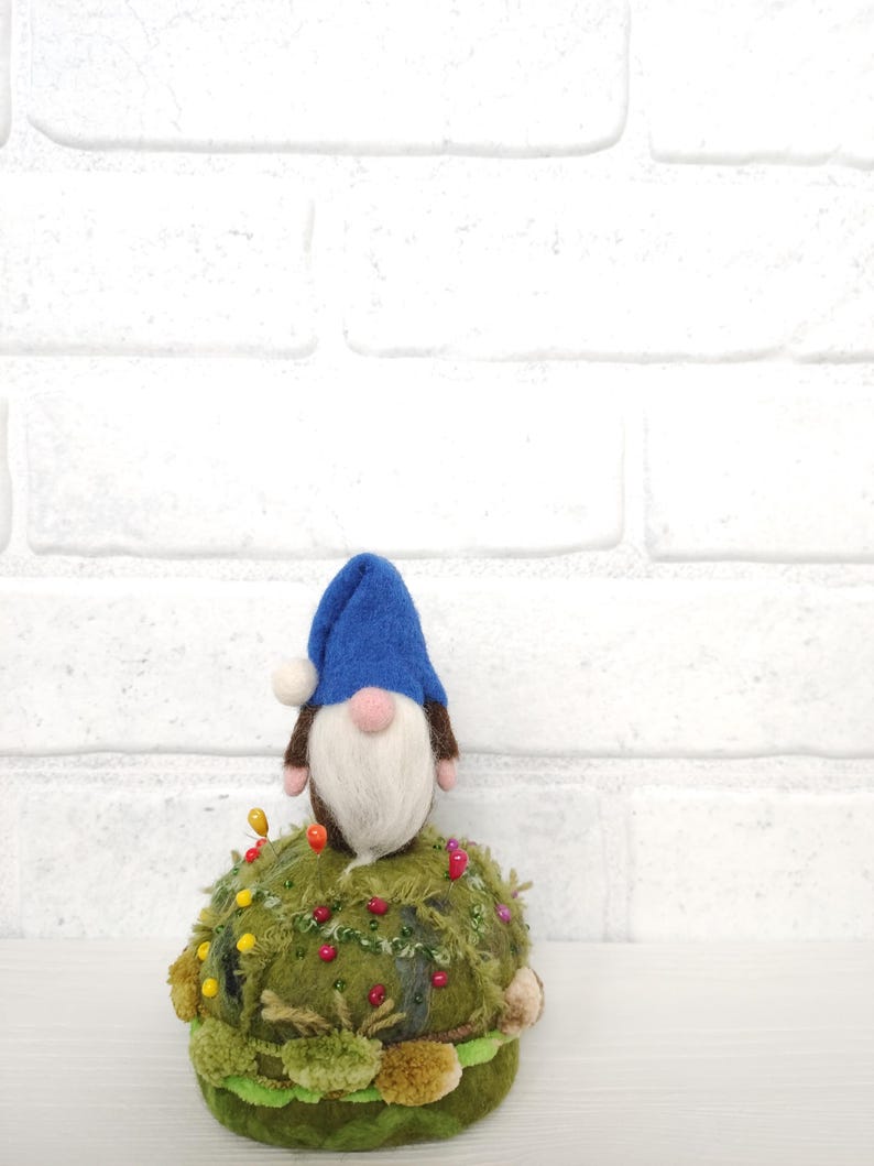 Puede incluir: Un gnomo de lana fieltrada con un gorro azul y barba blanca se asienta sobre un acerico redondo de color verde musgo. El acerico est&aacute; decorado con varios alfileres de colores, peque&ntilde;as cuentas y capas de tela texturizada.