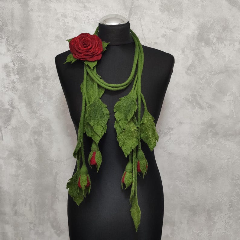 May include: A long felt necklace with a red rose and green leaves. The necklace is made of felt and has a long, flowing design. It is perfect for adding a touch of elegance to any outfit.