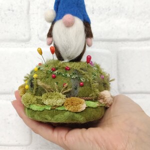 Puede incluir: Un alfiletero con un gnomo de fieltro con sombrero azul, barba blanca y nariz rosa. La base es un mont&iacute;culo verde texturizado decorado con peque&ntilde;os pompones, hilo verde y varios alfileres de costura de colores insertados en la superficie.