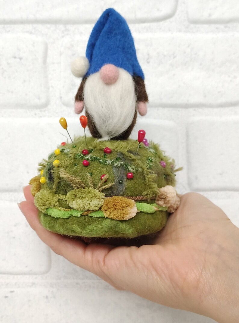 Puede incluir: Un alfiletero con un gnomo de fieltro con sombrero azul, barba blanca y nariz rosa. La base es un mont&iacute;culo verde texturizado decorado con peque&ntilde;os pompones, hilo verde y varios alfileres de costura de colores insertados en la superficie.
