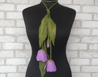 May include: A green felt scarf with a floral design. The scarf features large green leaves and three purple tulip-shaped flowers. The scarf is displayed on a black mannequin against a white brick wall.