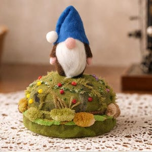 Puede incluir: Un acerico redondo de fieltro verde oliva decorado con peque&ntilde;os pompones coloridos y alfileres, rematado con una figura de gnomo de fieltro de aguja que lleva un sombrero azul puntiagudo con un pomp&oacute;n blanco, barba blanca y nariz rosa.