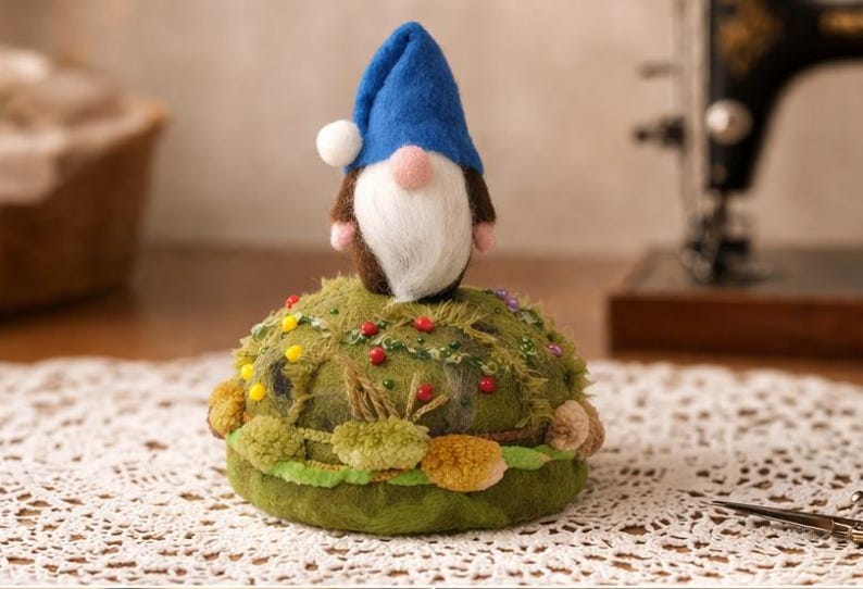 Puede incluir: Un acerico redondo de fieltro verde oliva decorado con peque&ntilde;os pompones coloridos y alfileres, rematado con una figura de gnomo de fieltro de aguja que lleva un sombrero azul puntiagudo con un pomp&oacute;n blanco, barba blanca y nariz rosa.