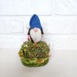 Puede incluir: Un alfiletero con un gnomo de fieltro con gorro azul, barba blanca y nariz rosa, posado sobre una base redonda con textura de musgo. La base verde est&aacute; decorada con varios alfileres de costura de colores y peque&ntilde;os detalles de tela.