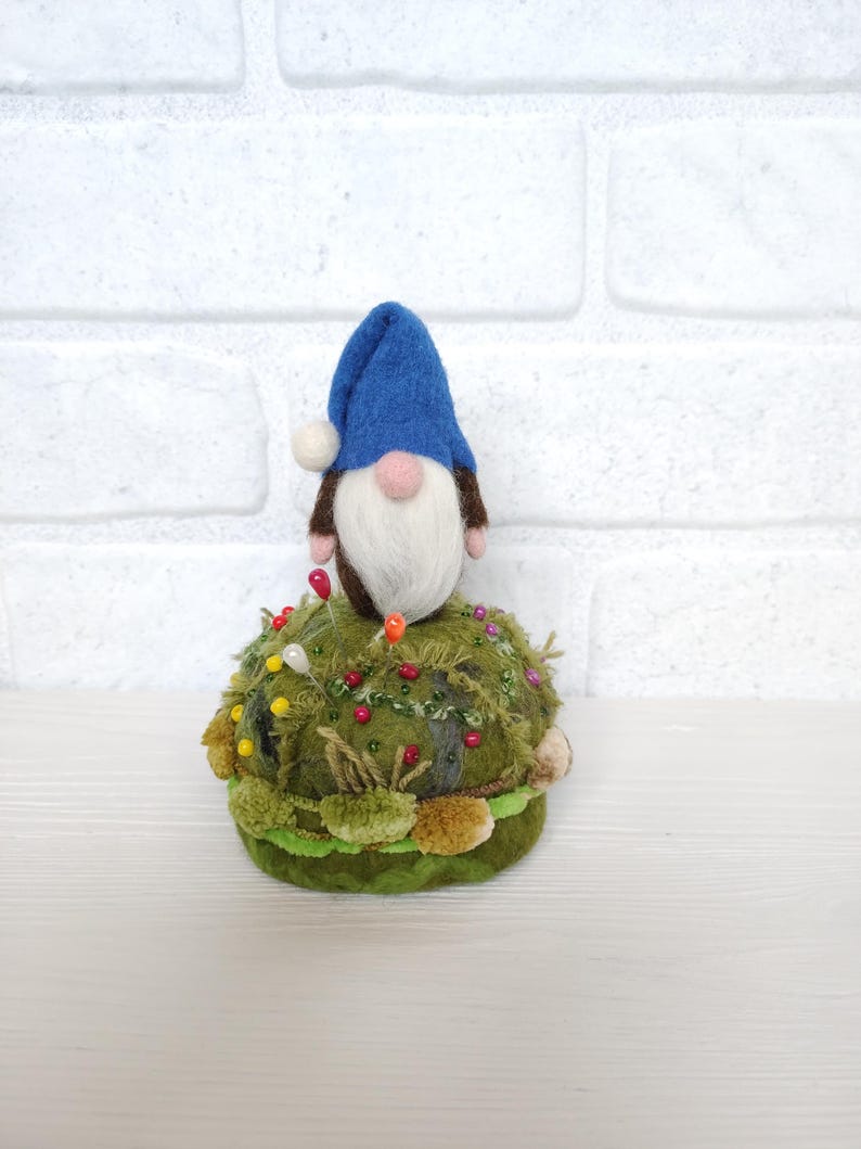 Puede incluir: Un alfiletero con un gnomo de fieltro con gorro azul, barba blanca y nariz rosa, posado sobre una base redonda con textura de musgo. La base verde est&aacute; decorada con varios alfileres de costura de colores y peque&ntilde;os detalles de tela.