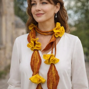 Puede incluir: Una bufanda larga de fieltro que presenta una mezcla de fibras de color naranja óxido intenso y amarillo vibrante. La bufanda está adornada con detalles tridimensionales en forma de flores con volantes y segmentos alargados.