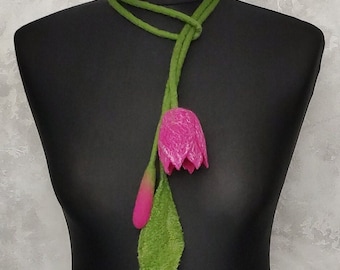 May include: A handmade felt necklace featuring a vibrant pink tulip flower and bud, with a green stem and leaf. The necklace is displayed on a black mannequin, showcasing the floral design.