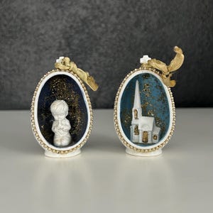 May include: Two white ceramic egg-shaped ornaments with gold trim. One ornament features a small white figurine of a boy praying inside. The other ornament features a small white church inside. Both ornaments have a blue background with gold glitter.