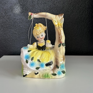 Puede incluir: Una figurita de cerámica de una niña con un vestido amarillo sentada en un columpio. El columpio está sujeto a un tronco de árbol con una mariposa en él. La niña tiene el pelo rubio y los ojos azules. La figurita está decorada con flores azules y hojas verdes.