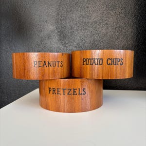 May include: Three wooden bowls with labels for snacks. The bowls are stacked on top of each other. The top bowl is labeled "Potato Chips", the middle bowl is labeled "Pretzels", and the bottom bowl is labeled "Peanuts".