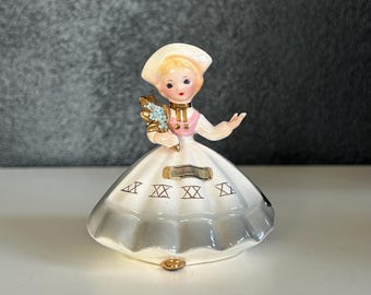 May include: A porcelain figurine of a woman wearing a white dress with a gray skirt and a pink bodice. She is holding a bouquet of blue flowers and has a white hat on her head. The figurine is labeled "Sweden".