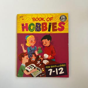 Puede incluir: Un libro vintage titulado "Book of Hobbies" con una portada amarilla e ilustraciones coloridas de niños que realizan diferentes pasatiempos. El libro es para niños y niñas de 7 a 12 años y tiene un sello de aprobación para niños.