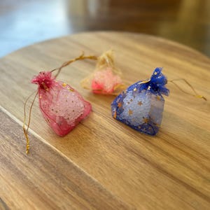 May include: Three small, sheer fabric bags filled with white, translucent beads. The bags are tied with gold string and have different colored patterns: pink, blue, and white.