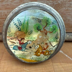 May include: A round, antique silver-toned compact mirror with a hand-painted scene of figures on horseback in a landscape. The artwork features vibrant colors and intricate details, set against a gold background.