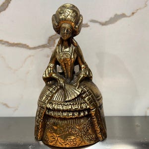 May include: A gold-toned brass bell shaped like a woman in a long dress and a large hat. The bell has intricate details and a floral design on the base.