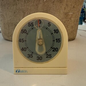 May include: A vintage Oster kitchen timer with a cream-colored body and a circular dial. The dial features black numbers on a light blue background, with a red indicator. The timer is set to approximately 55 minutes.