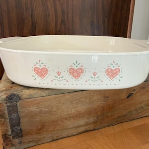 May include: A rectangular off-white ceramic baking dish featuring a repeating pattern of three pink hearts with white dots, interspersed with small green floral sprigs and dots along the front side, complete with integrated side handles.