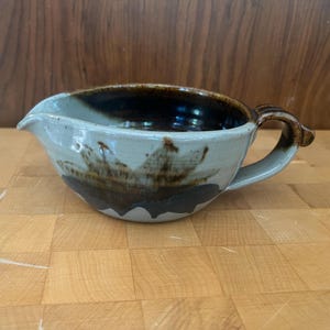 May include: A handmade ceramic mixing bowl with a brown and gray glaze. The bowl has a spout and a handle.
