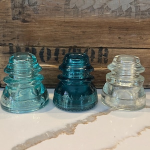May include: Three vintage glass insulators, one clear and two in shades of blue-green. The insulators are stacked in a pyramid shape and have a textured surface.