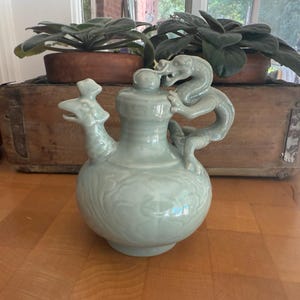 May include: A celadon-colored ceramic pitcher with a dragon handle and a bird-like spout. The pitcher has a bulbous body with embossed floral patterns. The background includes a wooden box with plants and a wooden table.
