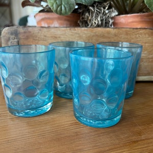 May include: Four light blue glass tumblers with a textured, bubble-like design. The glasses are arranged in a row on a wooden surface.